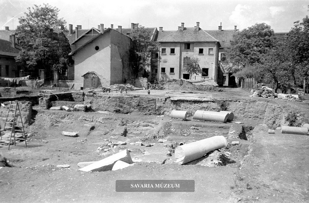 Az Iseum ásatása délről, kőfaragványokkal és a szentély udvarára dőlt in situ gránitoszlop maradványokkal. A háttérben a Thököly utca épületei láthatóak.